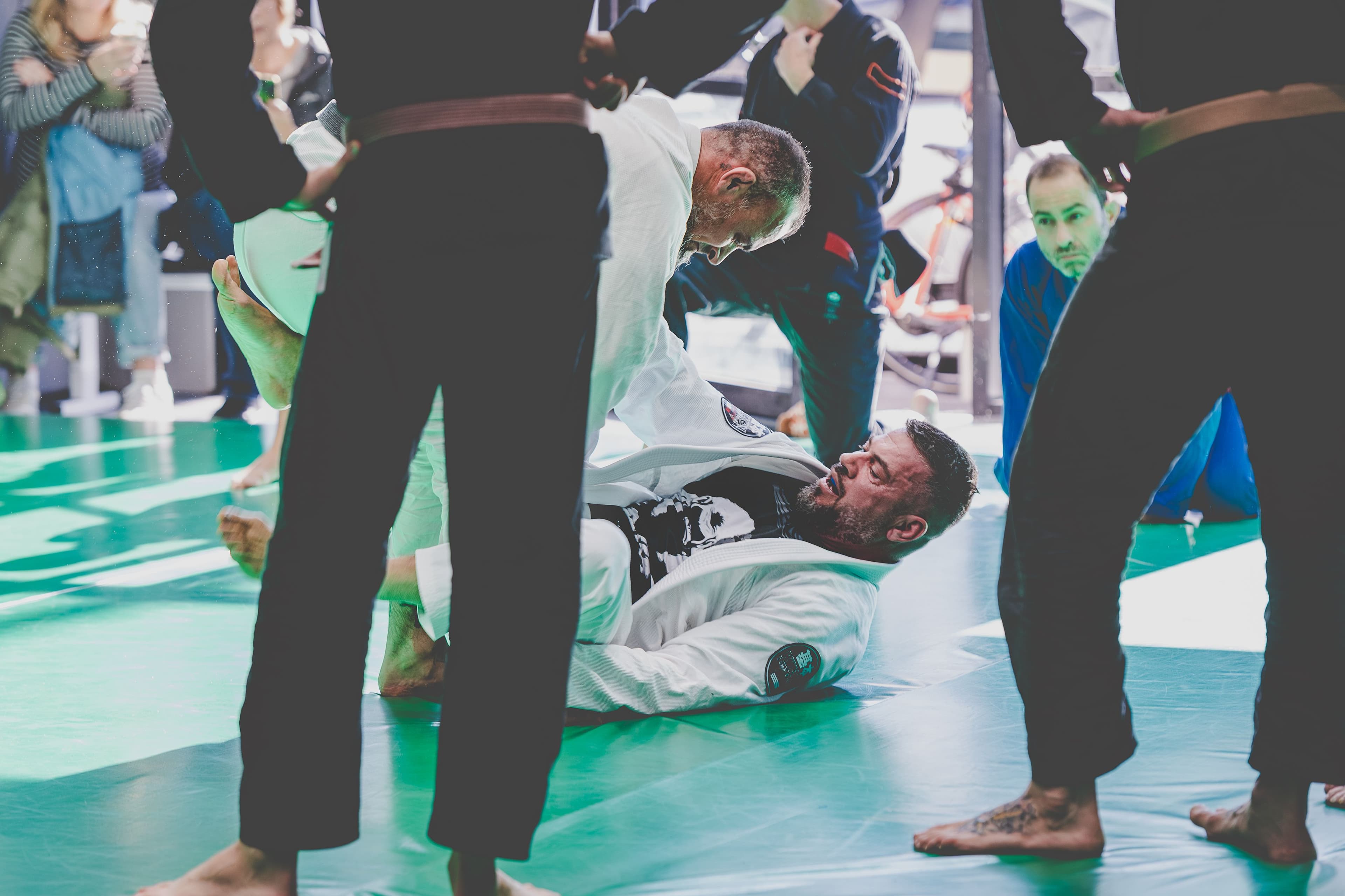 Two Brazilian Jiu-Jitsu practitioners spar on the mat during training, one on his back in a white gi controlling his opponent’s posture. Several teammates in gis stand around watching intently on the green mat under natural light.