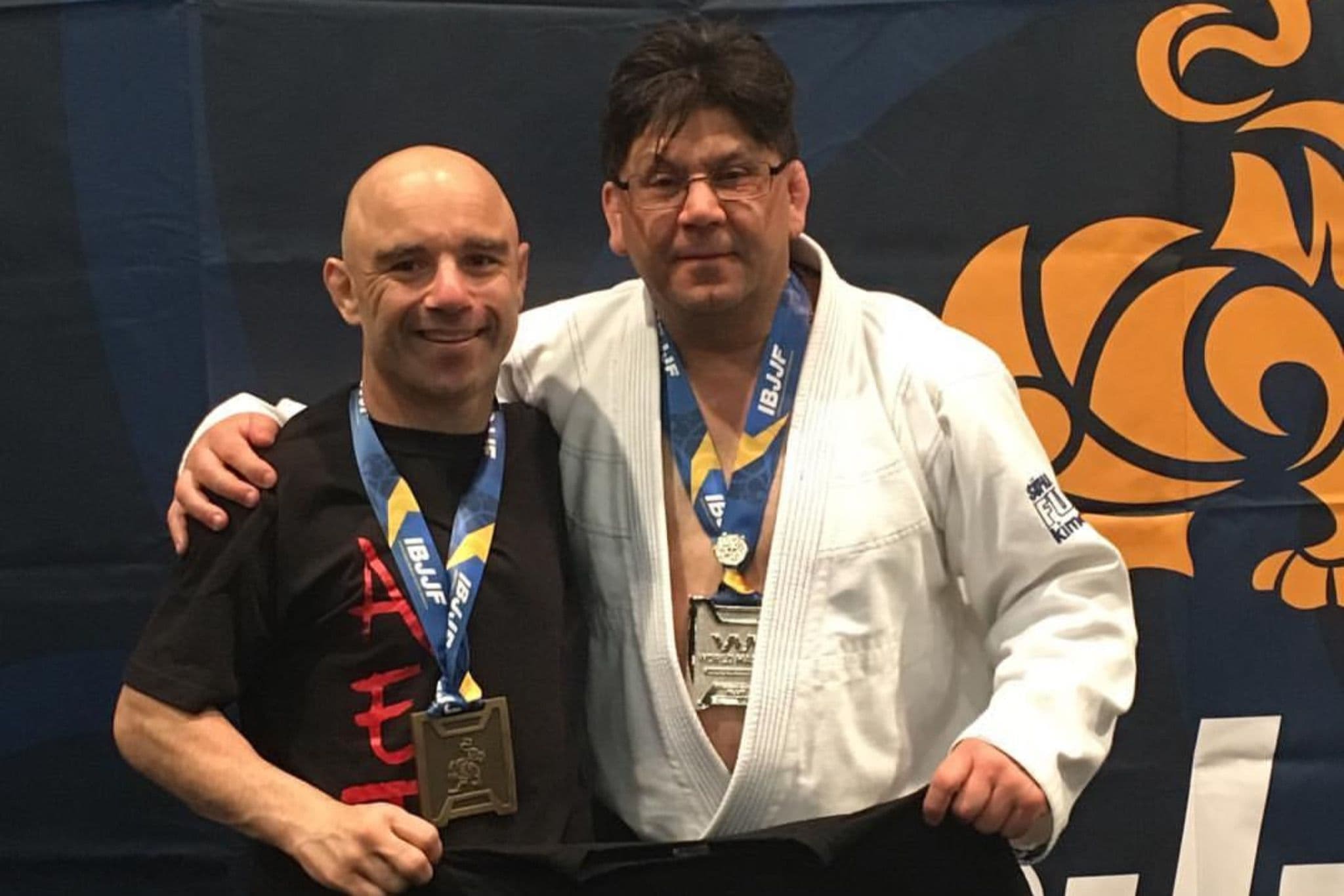 Two adult men standing side by side in front of a Brazilian jiu-jitsu competition backdrop, smiling and wearing medals on blue ribbons. One man is in a black T-shirt, the other is wearing a white gi. They are holding up a black shirt with a red graphic together.