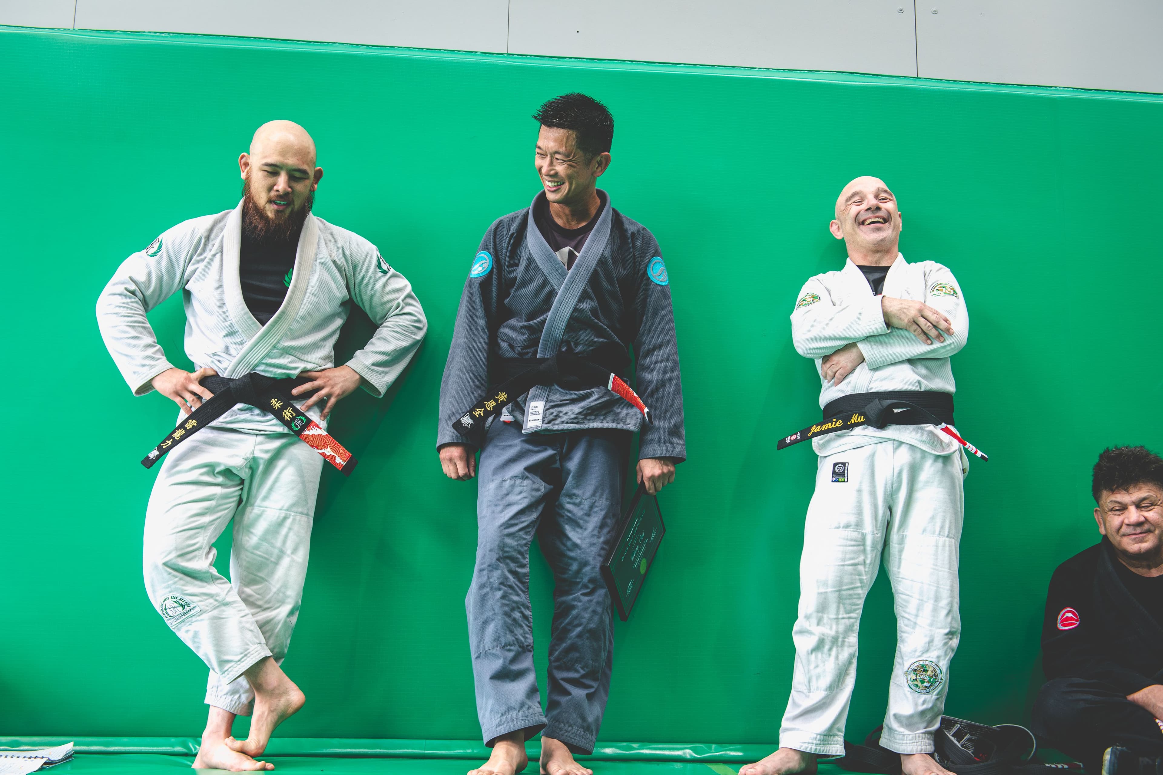 Three Brazilian Jiu-Jitsu black belts stand relaxed against a green padded wall, smiling and chatting after a grading ceremony, with others seated nearby.