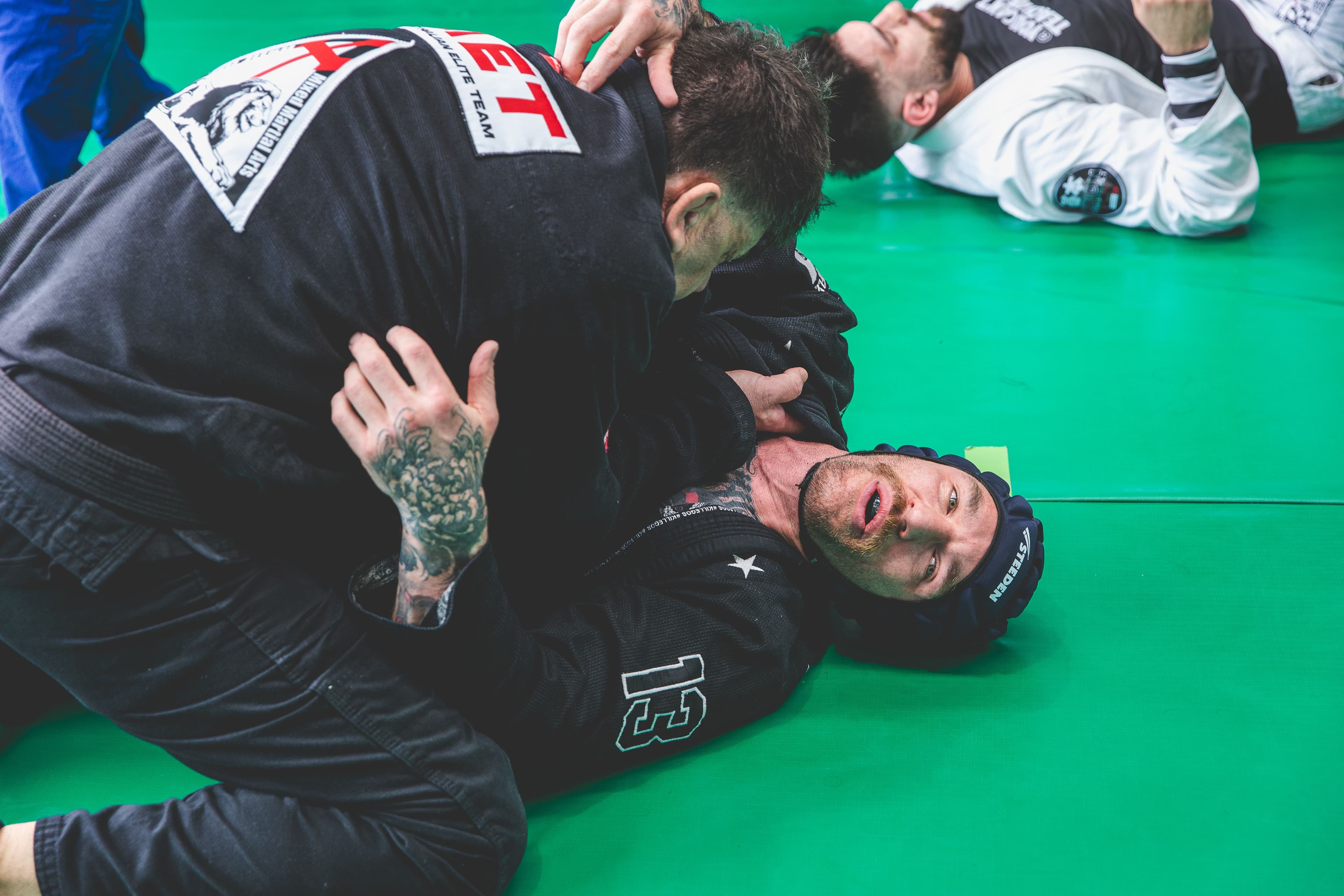 Two Brazilian Jiu-Jitsu practitioners roll on green mats, with one applying top pressure while the other looks toward the camera; additional students train in the background.