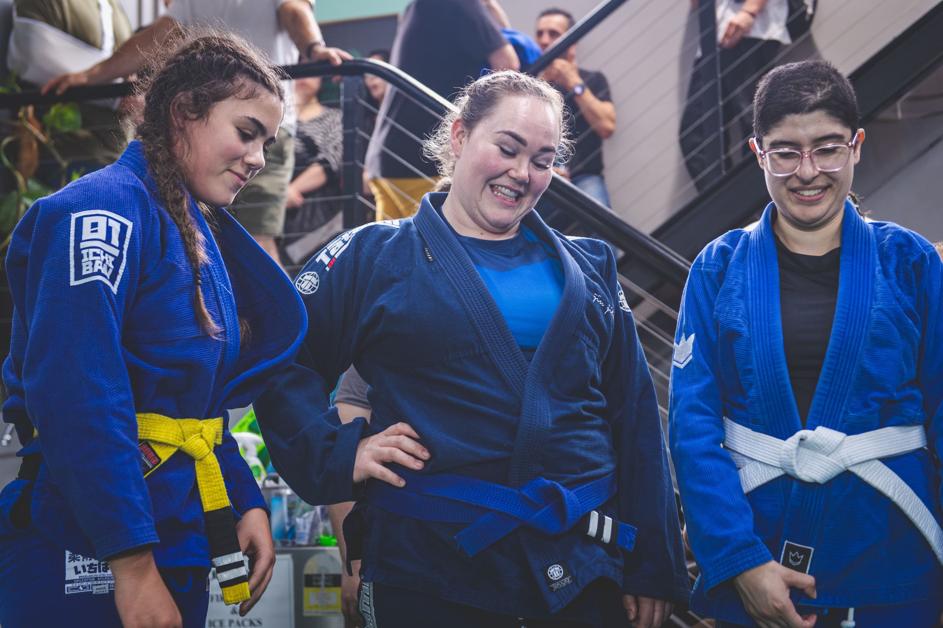 Three women in blue Brazilian Jiu-Jitsu gi uniforms smiling during a belt grading ceremony inside a martial arts academy, celebrating promotion and community achievement.