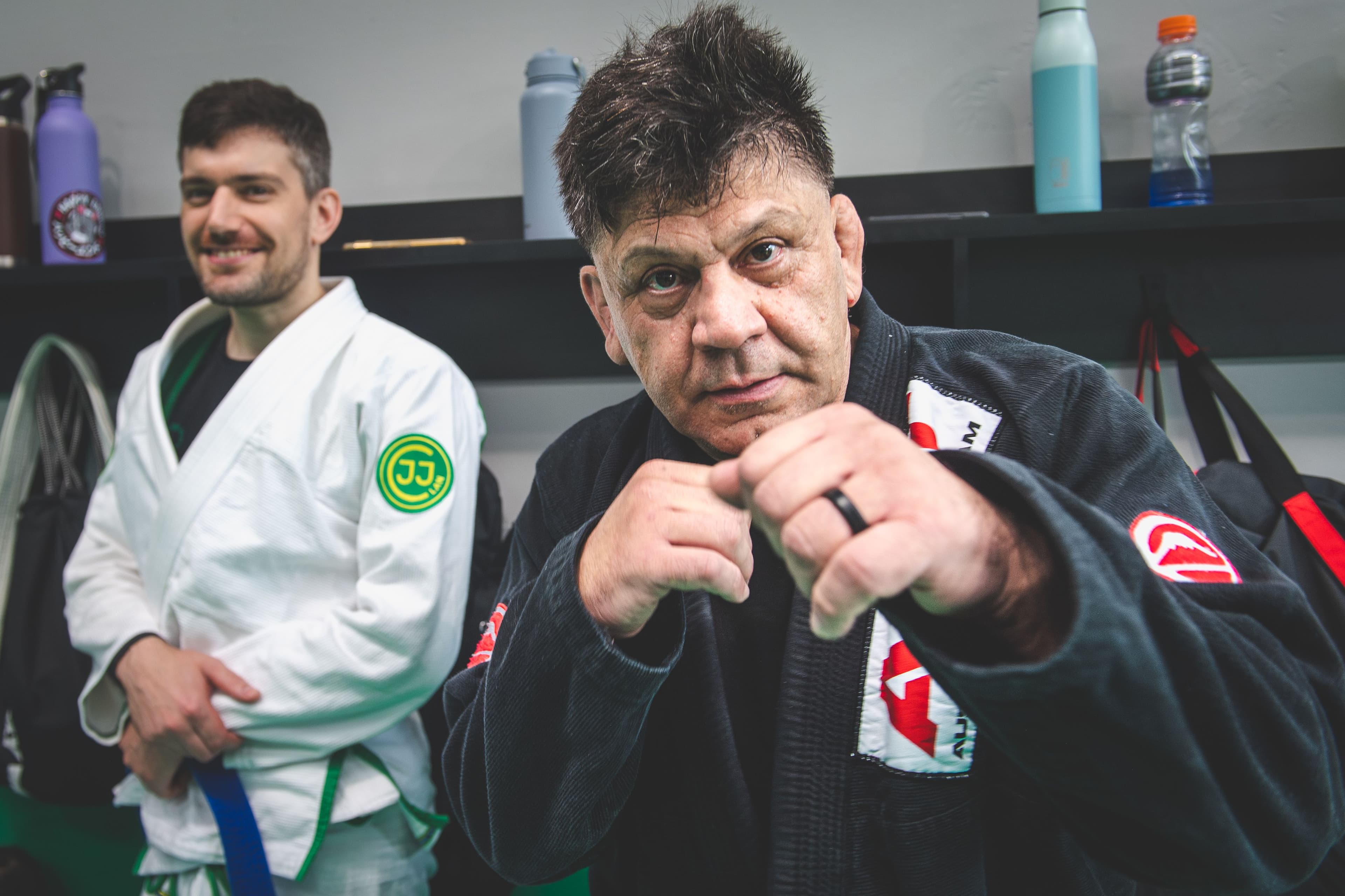 An older Brazilian Jiu-Jitsu practitioner in a black gi poses playfully with his fists raised, demonstrating confidence, energy, and enthusiasm for training.
