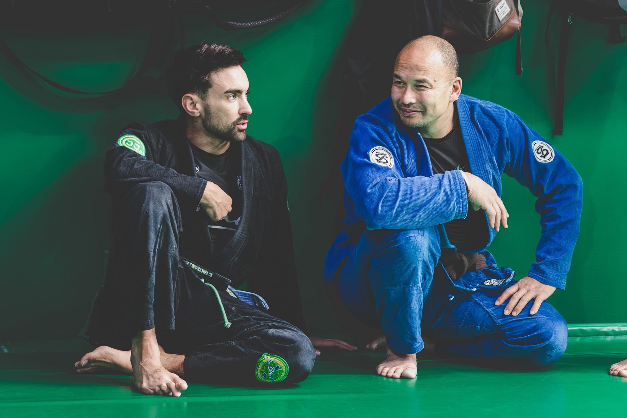 Two Brazilian Jiu-Jitsu practitioners sit on green mats mid-conversation, wearing gis (one black, one blue). They face each other with relaxed, friendly expressions, suggesting a break during training or a casual discussion between rounds.