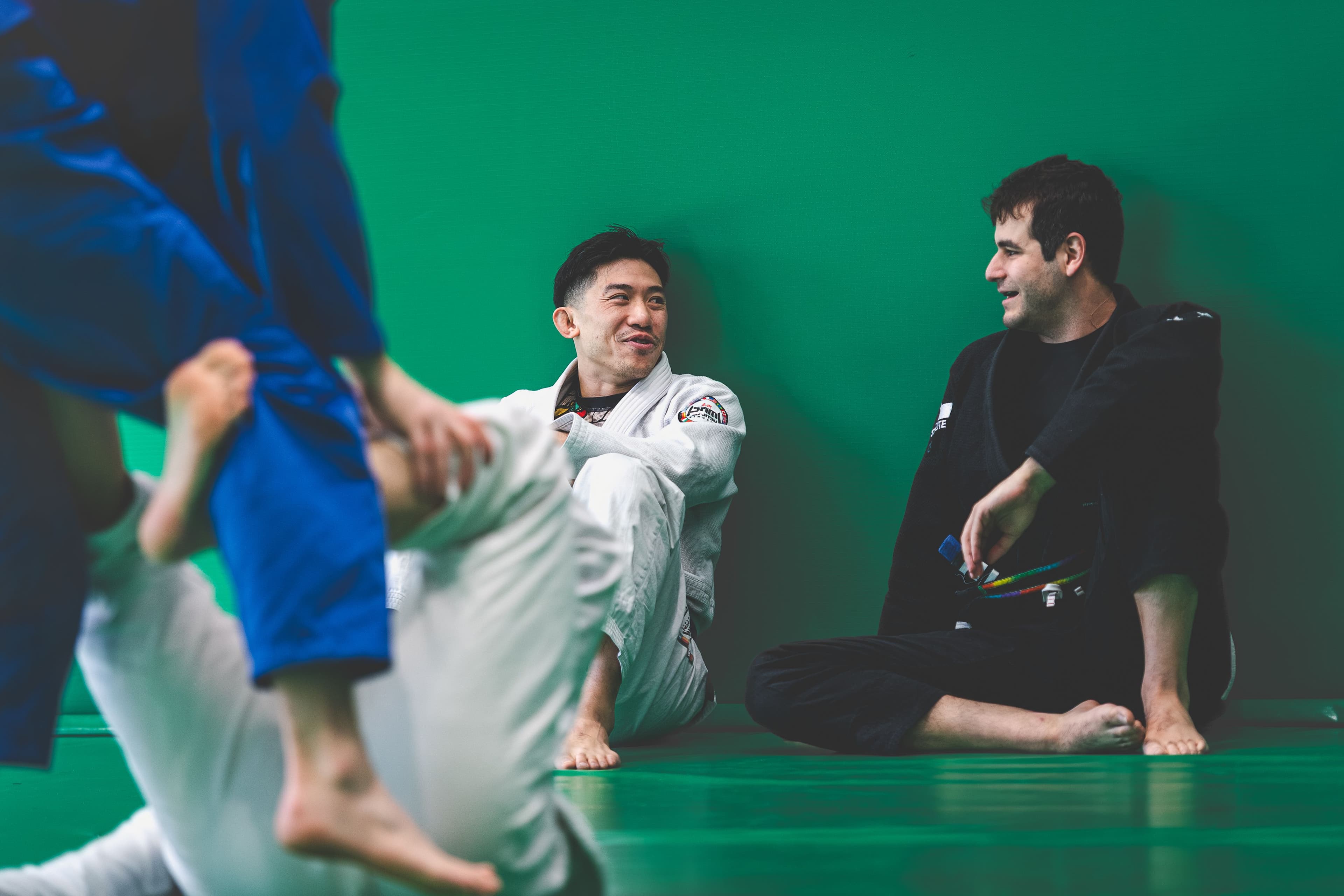 Two Brazilian Jiu-Jitsu practitioners sit relaxed on the edge of green mats, smiling and chatting after training. Both wear gis; one white, one black. While others roll in the background, creating a friendly, informal atmosphere on the mat.
