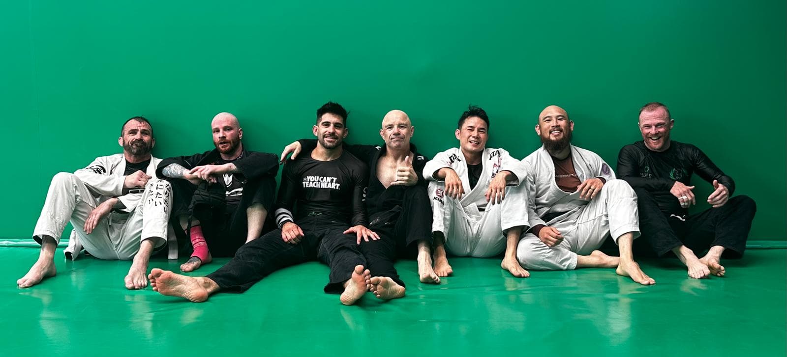 A group of adult Brazilian jiu-jitsu practitioners sitting barefoot on green mats, resting shoulder to shoulder after training. Some are wearing white gis and others black rash guards, smiling and relaxed against a green wall.
