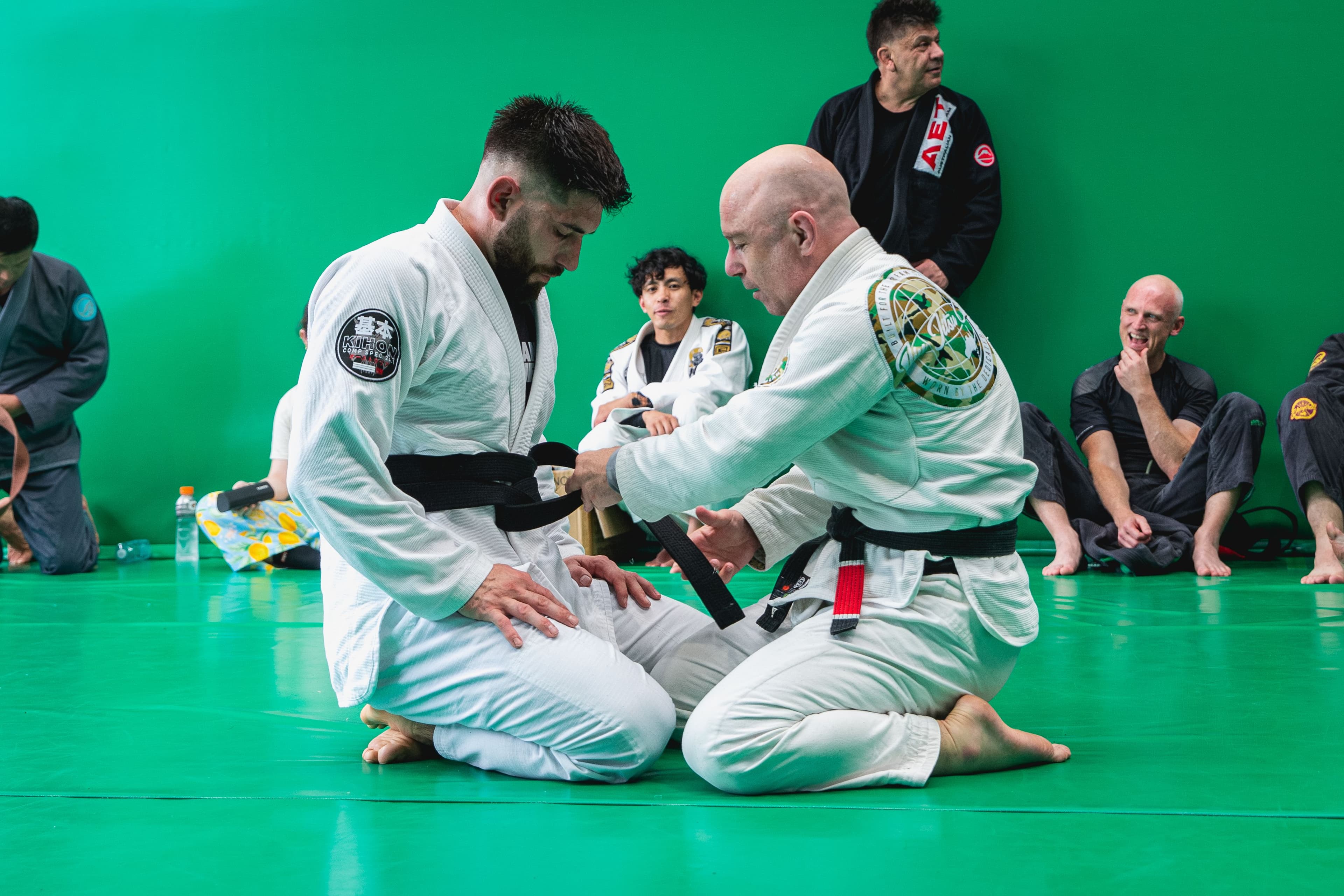 A Brazilian Jiu-Jitsu instructor kneels on green mats while tying a black belt around a kneeling student in a white gi, as other students watch from behind.
